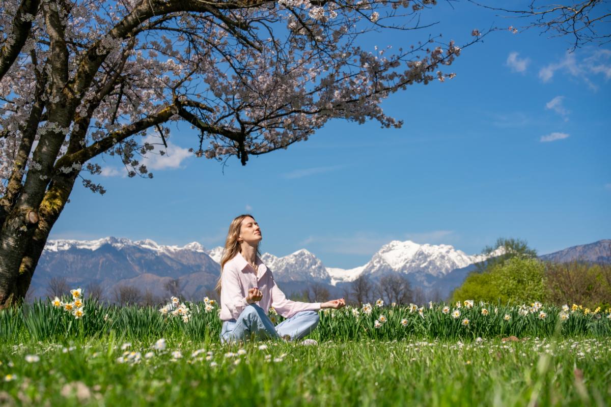 Young woman meditating under a cherry blossom tree with snowy mountains in the background enjoying a peaceful spring day. Digital detox Freedom and Spring vibes