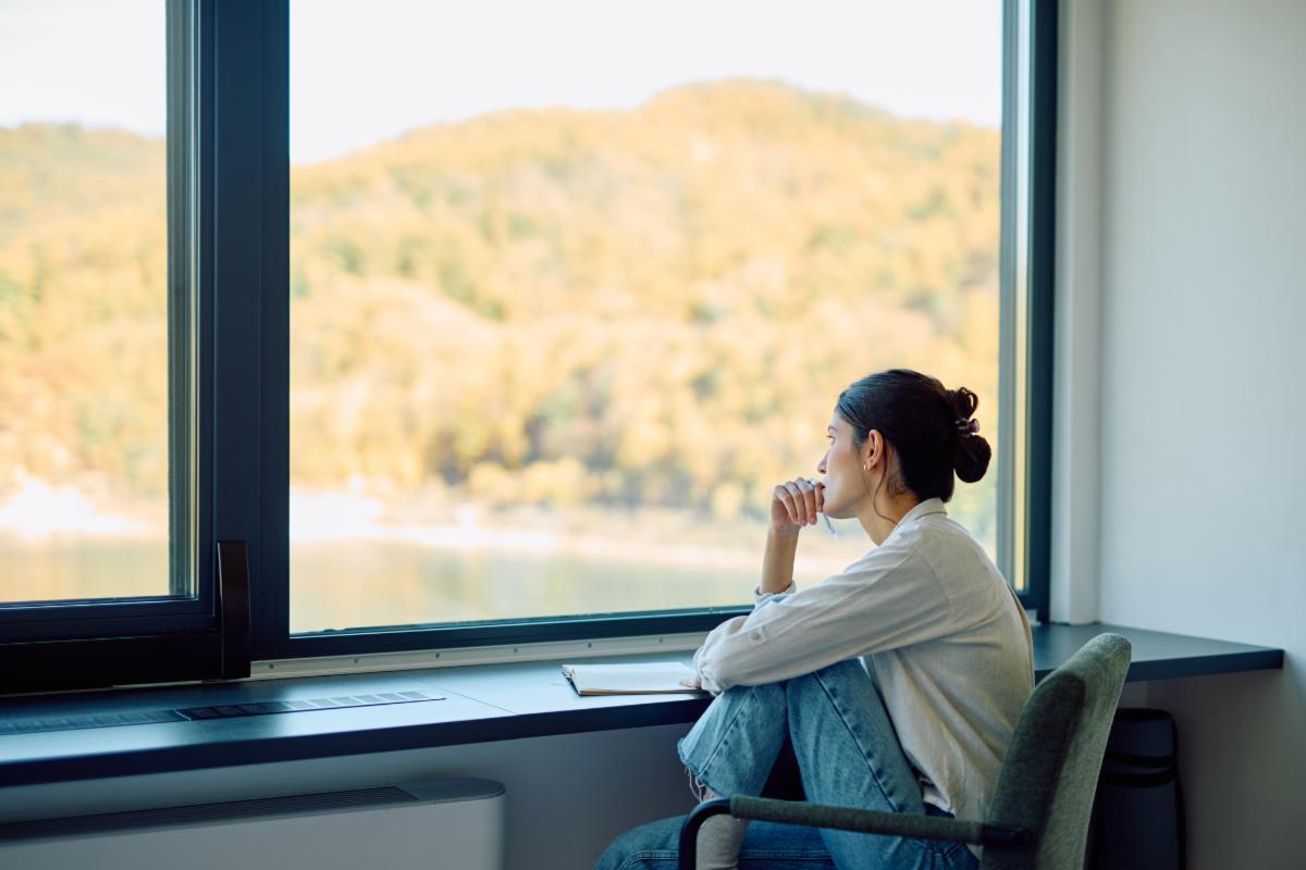 Young woman sitting by a large window looking out at the autumn landscape and lake thinking or contemplating