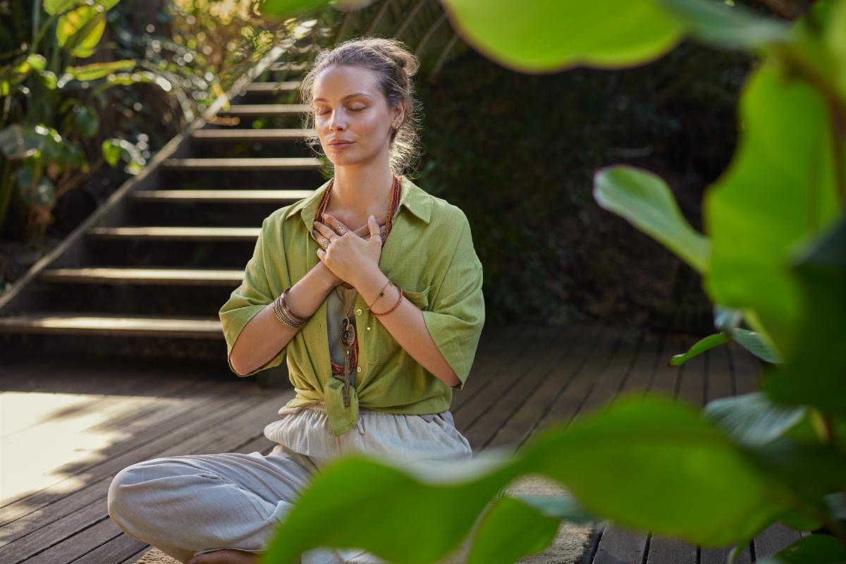 Peaceful woman sits cross-legged in a meditative state hands over her heart expressing gratitude and mindfulness in a lush. Girl connects with her emotions during a reflective moment embracing inner peace. Calm woman practices a heart-centered meditation c