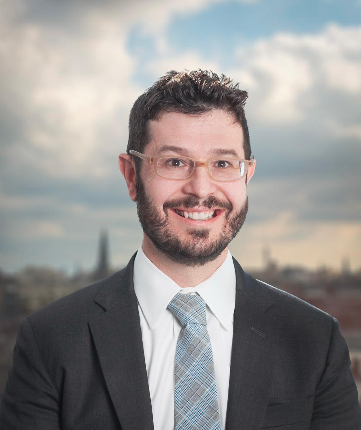 Profile photo of Todd, wearing a light blue plaid tie and grey suit. He is wearing glasses and has a brown beard. 