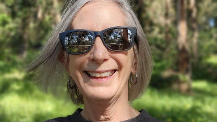 Headshot of Cynthia Rice. She is a white woman with a silvery bob hairstyle. In this photo she is wearing sunglasses and smiling at the camera with greenery in the background