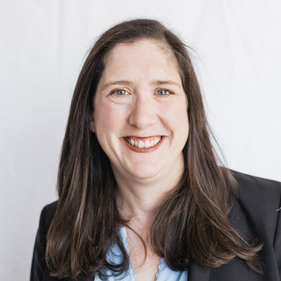 Headshot of Jennifer Klar. She is smiling at the camera and has long brown hair parted at the right side. She is wearing a blue blouse with a suit jacket over.