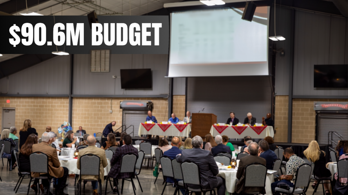 Attendees seated at round tables during a public meeting, with panelists at the front of the room. Text overlay: â$90.6M Budget.â  