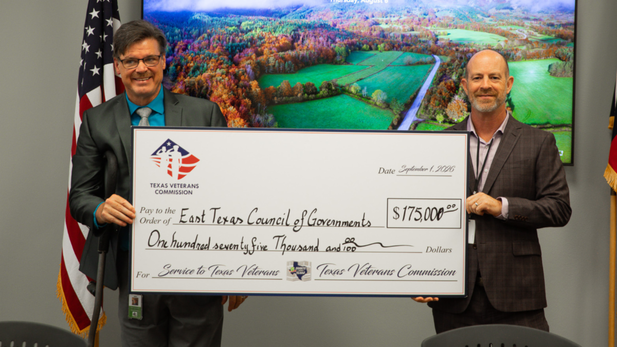 Two representatives hold a ceremonial $175,000 check from the Texas Veterans Commission to the East Texas Council of Governments for services to Texas veterans, standing between U.S. and Texas flags.  