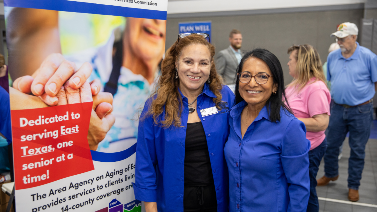Two Area Agency on Aging of East Texas staff members pose at an event booth promoting services for older adults. A banner reads, âDedicated to serving East Texas, one senior at a time!â