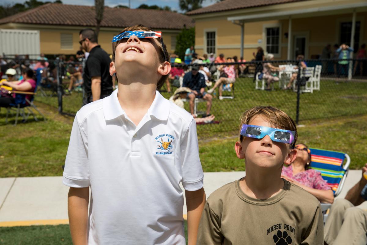Two boys wearing solar eclipse glasses look up at the sky.