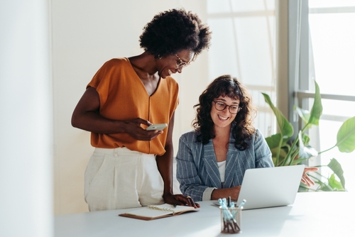 Happy professional women collaborating with enthusiasm in a modern office setting. Two female entrepreneurs using a laptop for a productive business meeting in a corporate workplace.
