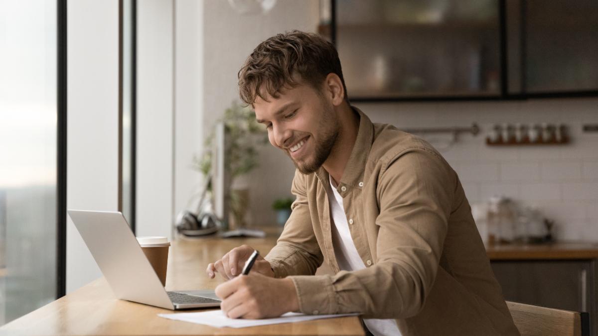 Wide banner panoramic view of smiling young Caucasian man sit at desk make notes working online on computer. Happy millennial male handwrite study distant on laptop in office. Education concept.