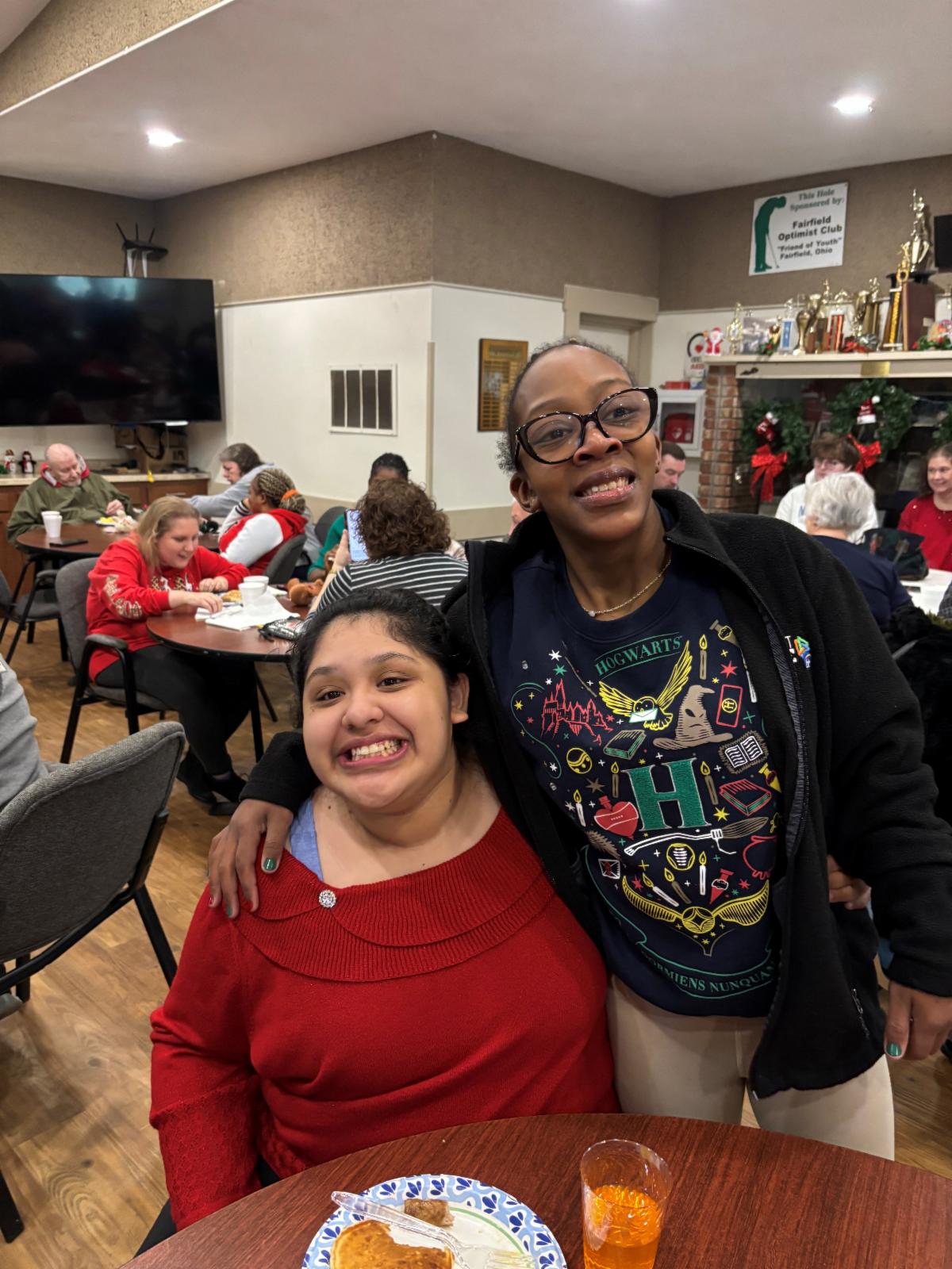 Two women smile at the camera. On the table in front of them is a glass of orange juice and a paper plate with pancakes. 