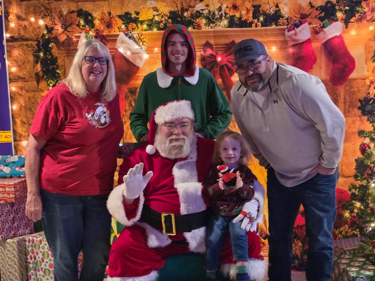 Santa holds a little boy. Behind him is an elf. A woman and a man stand on either side of Santa. They are all in front of a festive holiday backdrop.