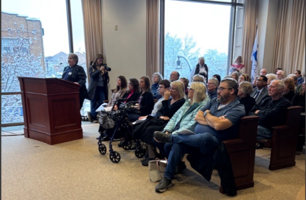 A woman speaks at a lectern. Behind her is a crowded room. 
