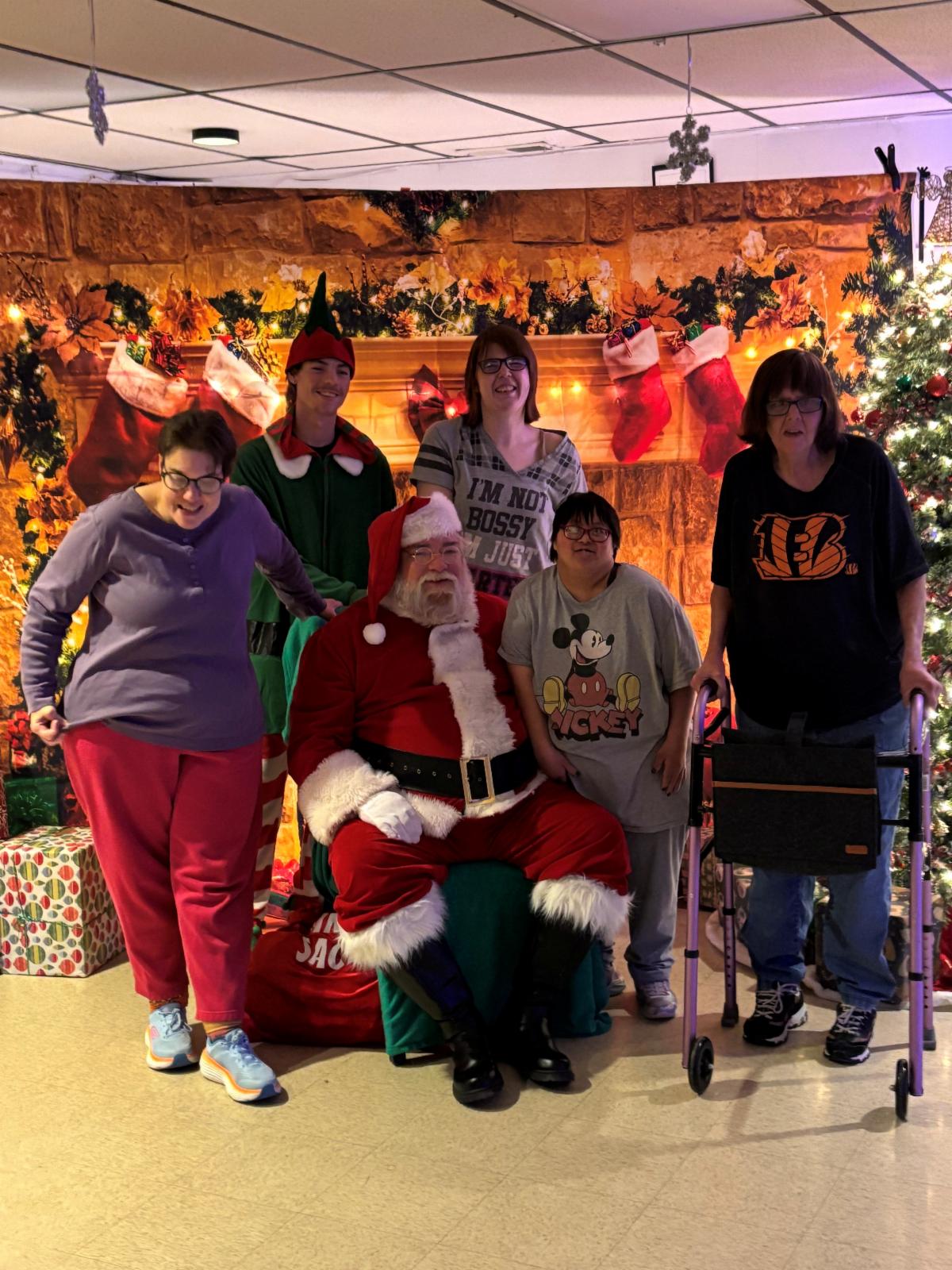 Santa sits among a group of people in front of a festive holiday backdrop. One person appears to have Down Syndrome. Another person uses a walker. 