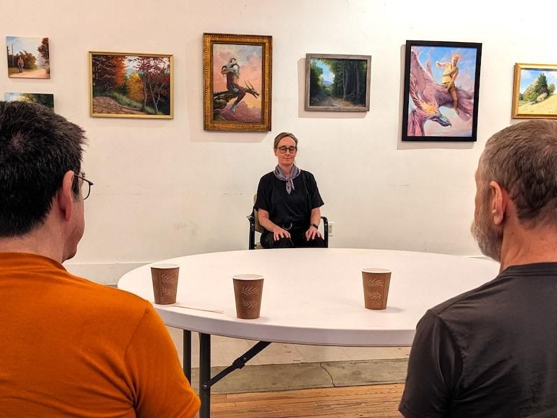 Alison Fox leading Rainbow Lounge group attendees in a guided meditation. Alison is sitting at one end of a table in front of the artwork with her eyes closed and two individuals are at the other side of the table.
