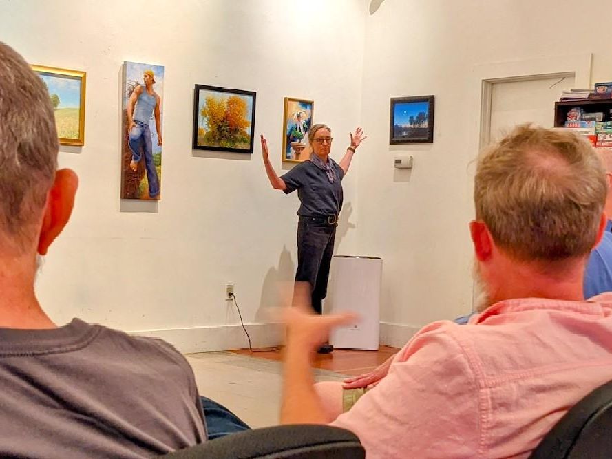 Alison Fox leading the attendees of the Rainbow Lounge in a discussion on the artwork by Ray Passer. Alison is standing in front of the art with individuals seated in front.