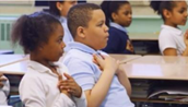 Photo of two children sitting side by side in a classroom, with both arms crossed on their chests and their hands on their hearts.