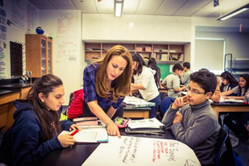 Photo of two middle school students sitting at a desk facing each other, with their teacher standing next to them pointing out work on the desk.