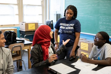 Photograph shows a classroom discussion with four students seated around a table. Students review papers and engage in conversation, with desks, computers, windows, and a chalkboard filled with writing visible in background.