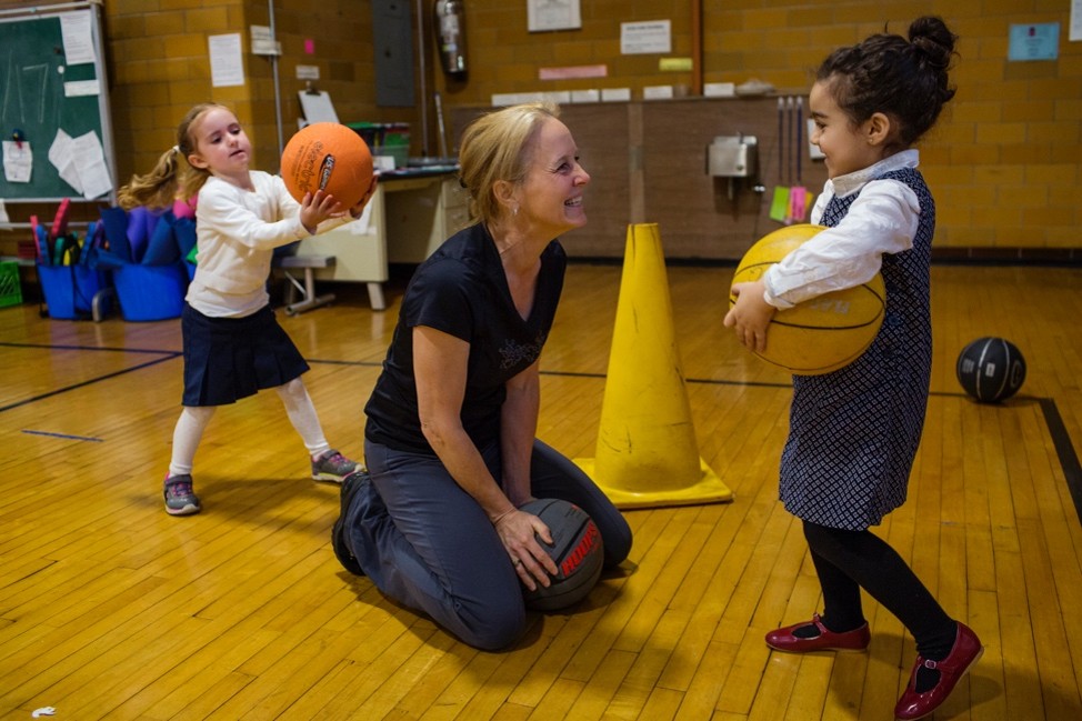 Photograph shows a gym basketball activity with an adult instructor kneeling beside a young girl holding a ball. In the background, another child prepares to pass an orange basketball.