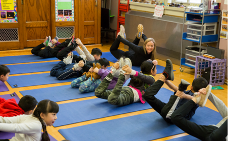 Photo of elementary students and their teacher in a classroom, lying stomach-down on yoga mats and doing a yoga pose.