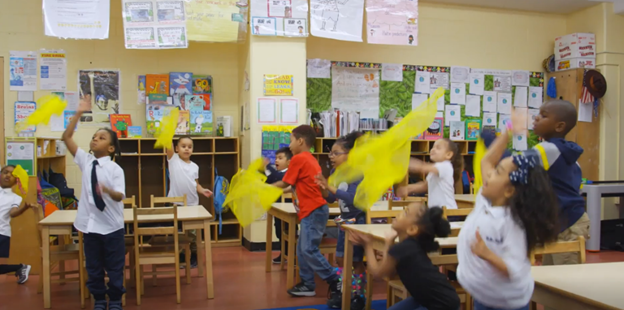 Photo of elementary school children standing up in a classroom around their desks and waving yellow scarves in the air.