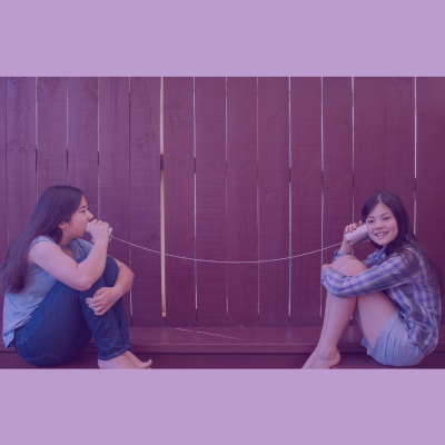 Two teenage girls sitting across from each other each holding a tin can connected by a string between them. One girl is talking into her can. The other girl has her can to her ear listening.