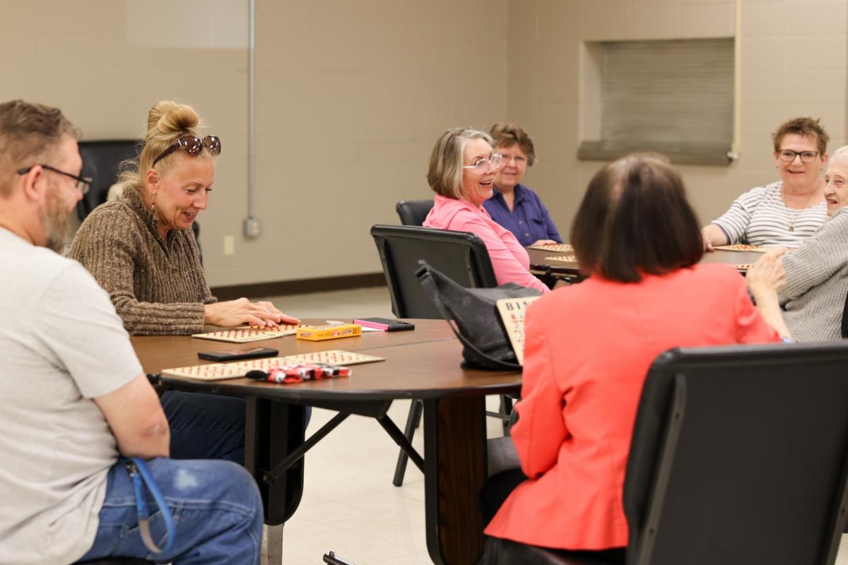 A group of adults sit around tables playing bingo at the De Soto Senior Center. Several players smile and talk while bingo cards, markers, and prizes sit on the tables.