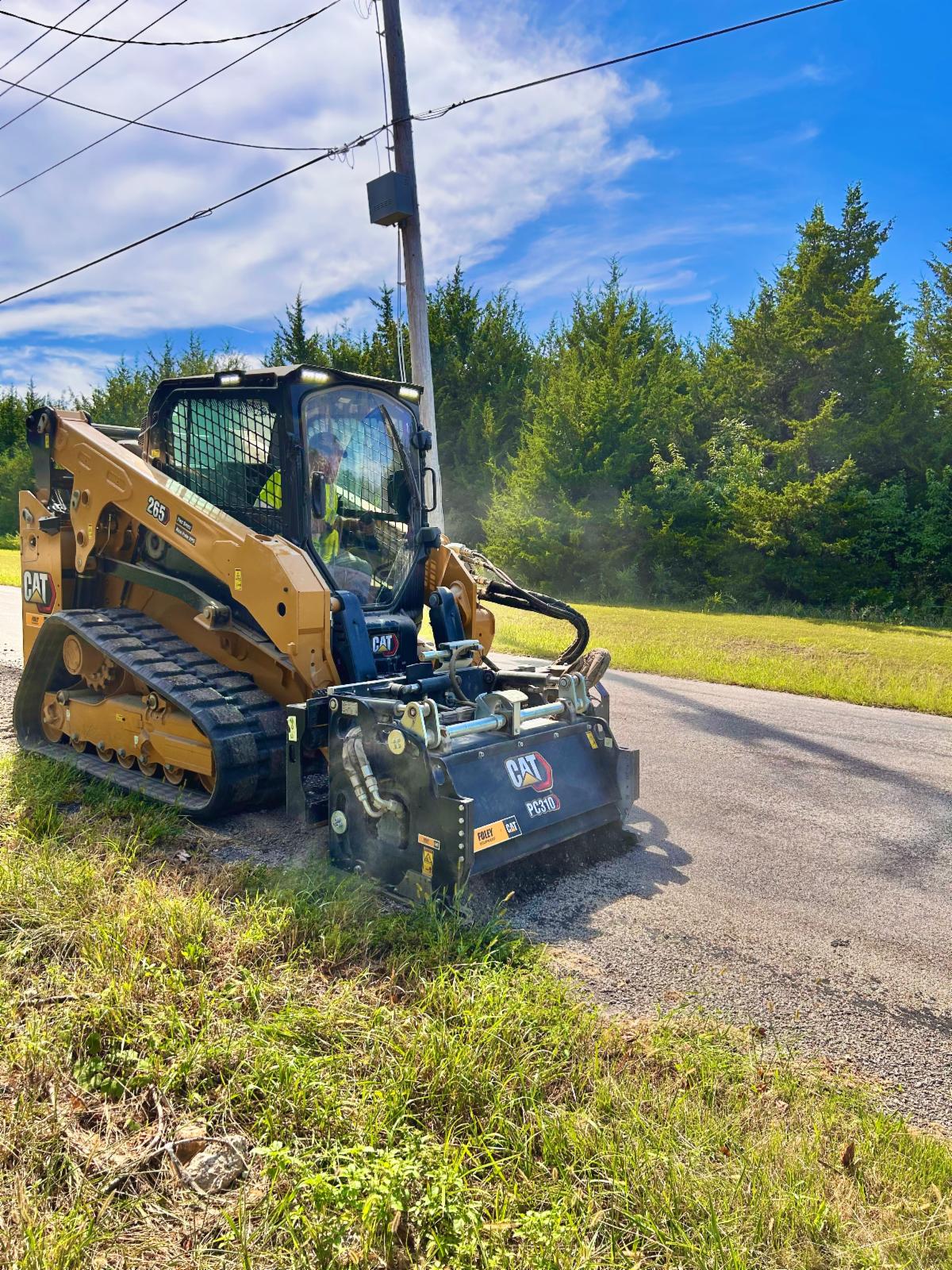 A man in a yellow construction shirt operates a milling machine along the edge of a roadway.