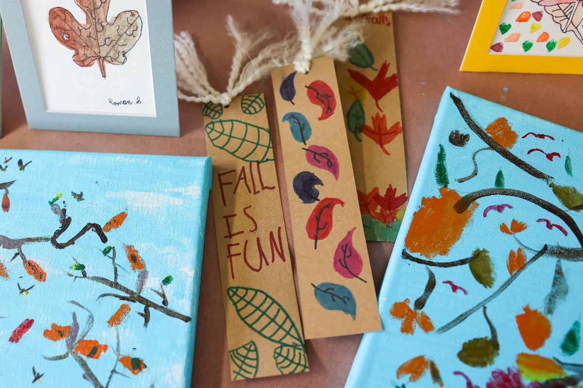 Fall-themed art projects done by kids are scattered across the table. In the center is a bookmark covered in leaves that reads, "Fall Is Fun."
