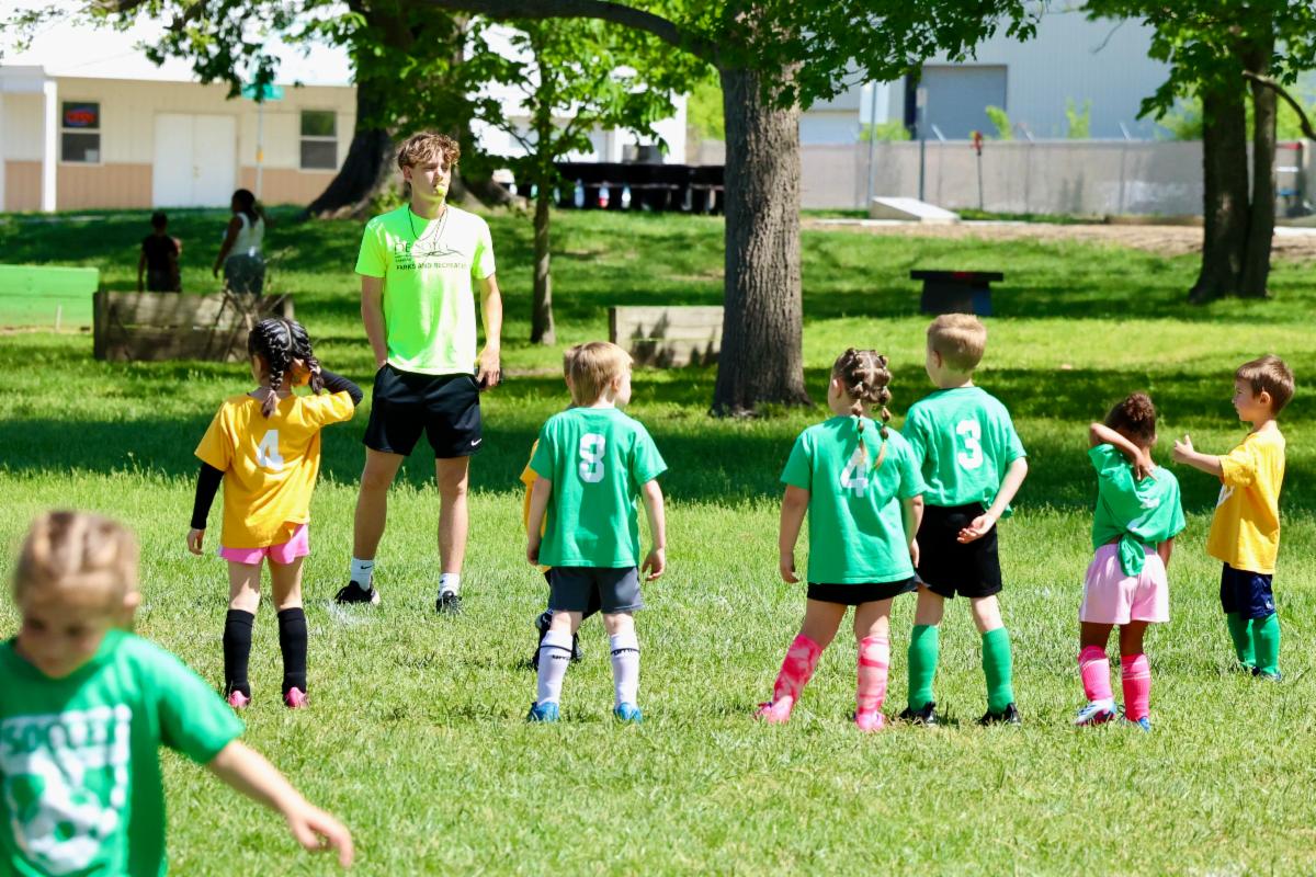 Young soccer players stand on a grassy field facing an official who has a whistle in his mouth and is wearing a De Soto Parks & Recreation shirt. 