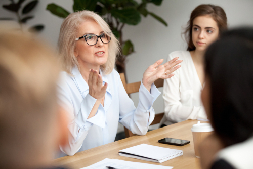 Attractive aged businesswoman_ teacher or mentor coach speaking to young people_ senior woman in glasses teaching audience at training seminar_ female business leader speaker talking at meeting