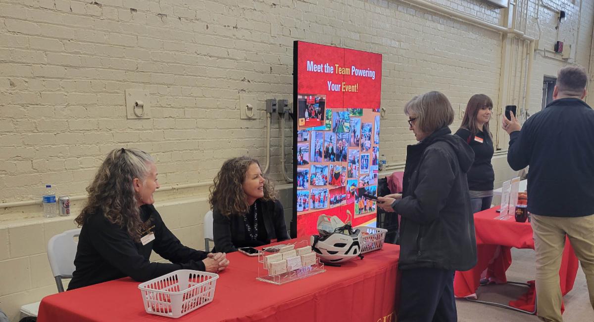 Two women sit at a draped conference table with giveaway gifts and candy while chatting with another woman standing across from them