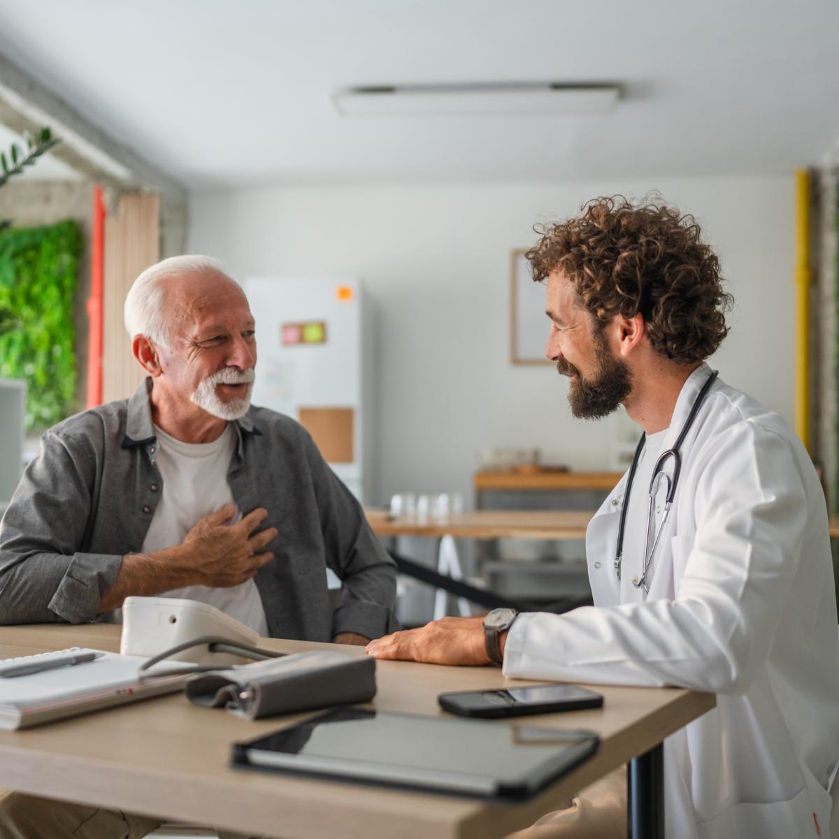 Adult doctor visiting a senior man patient and talking about healthcare treatment in a home medical office with blood pressure monitor pills notebook pen tablet and smart phones on the table