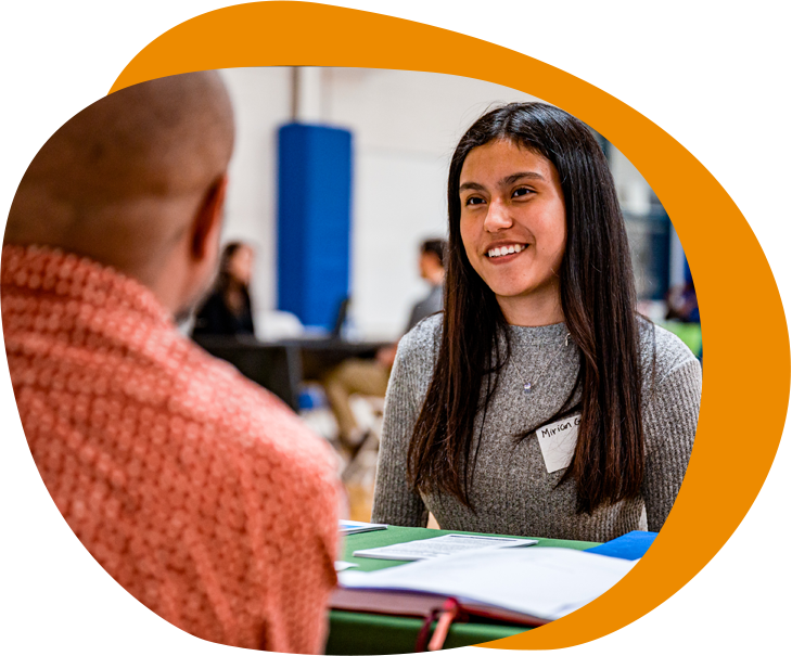 A young woman of color with long black hair and a grey sweater sits across from someone and speaks to them. 