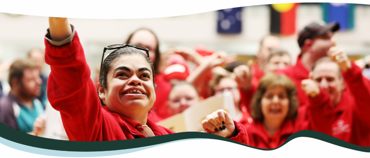 A young woman in a red jacket smiles and waves towards a crowd. A group of people all in red, standing behind her, wave to the crowd as well. 