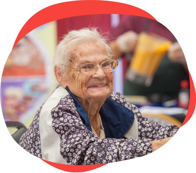 An older white woman wearing a white and purple jacket with short white hair and glasses sits and smiles at the camera.