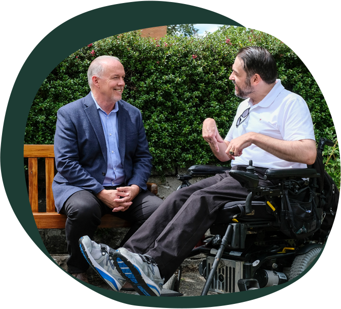 An older white man sits on a park bench, engaged in conversation with a younger white man using a wheelchair.