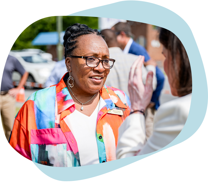 An older Black woman with glasses, a colorful shirt, and a braided hairstyle speaks to someone off-camera.