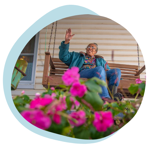 An older black woman sits on a porch swing smiling and waving to someone in the distance. 