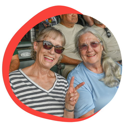 Two older white woman sitting together smile at the camera. One has short greying hair and sunglasses. She is wearing a striped shirt and holding up a peace sign. The other woman has longer white hair. She is wearing sunglasses and a blue shirt. 