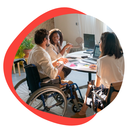 A group of individuals sits around a table discussing something. The main man in the photo is a wheelchair user. He is wearing a tan shirt. He has short black hair and a black beard. He is speaking to a black woman who is also wearing a tan shirt. She has shoulder length curly hair. 