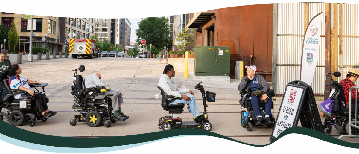 A number of individuals in wheel chairs are in line for something. The photo shows them spaced evenly a part while they wait on a city sidewalk.