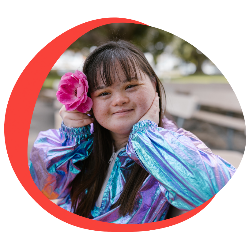A young girl with down syndrome sits and smiles at the camera. She has black long hair with bangs. She is wearing a multicolored iridescent jacket with a pink flower in her hair.