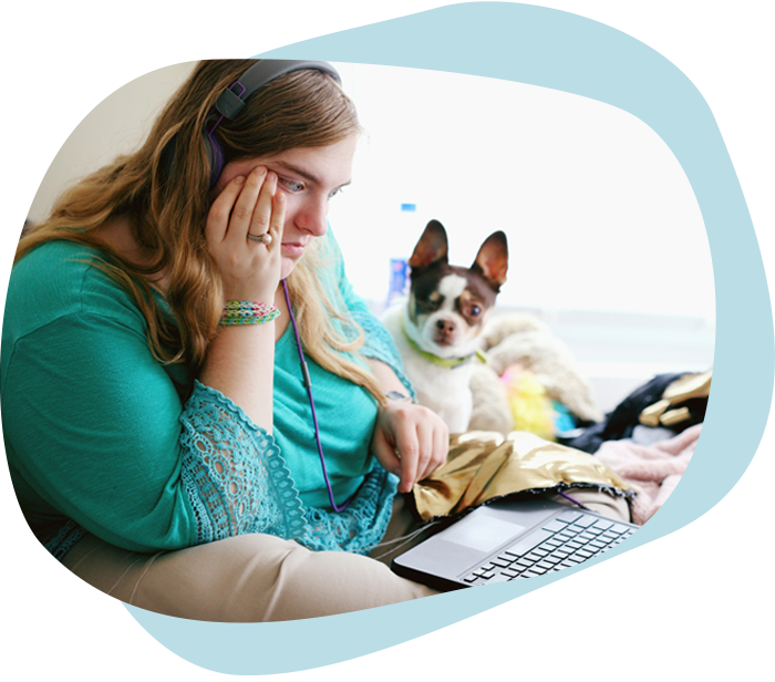 A young white woman with a blue shirt and headphones sits on her bed looking at her computer. A small dog sits next to her. 