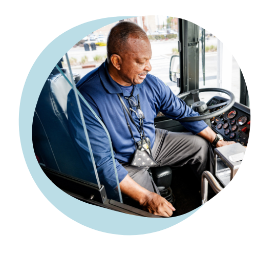 An older black man sits in the front seat of a public bus explaining the different bus controls of a new more accessible bus. 