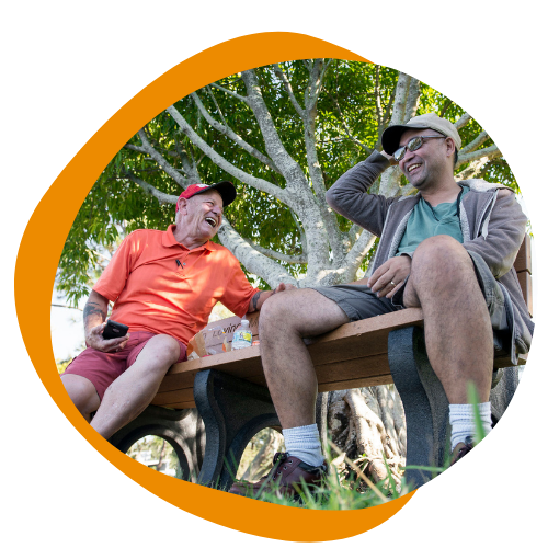 Two men sitting on a picnic table outside laughing and smiling at each other. 