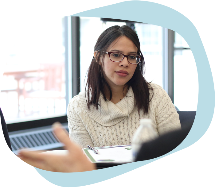 A young woman of color in glasses and a white sweater listens attentively to a speaker off-camera.