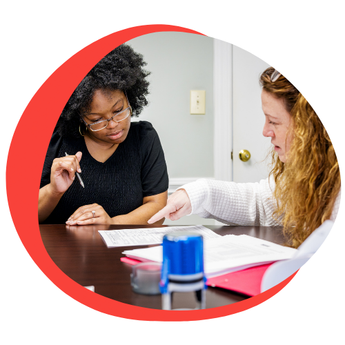 A young black woman with a black shirt and glasses sits at a table looking over a document. A young white woman with brown hair and a white jacket sits next to her and does the same. 
