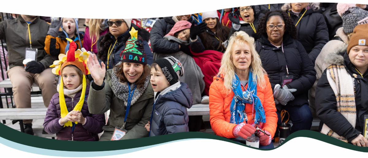 A group of people sitting and watching a winter parade. There is a mother with two children waving at the camera with a "holiday" hat on. A second blonde woman sits next to her smiling at the camera. 