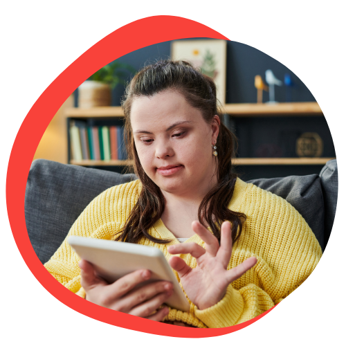 A young woman with Down syndrome sits and scrolls through an iPad. She is wearing a yellow sweater. She has brown hair that is pulled back on the top.