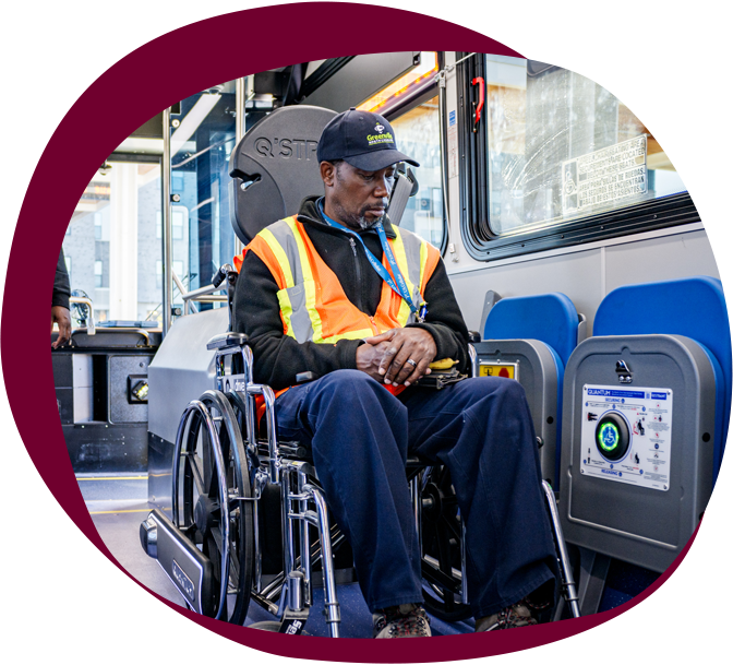 A middle aged black man wearing a cap and an orange vest sits in a wheelchair on a public bus. 